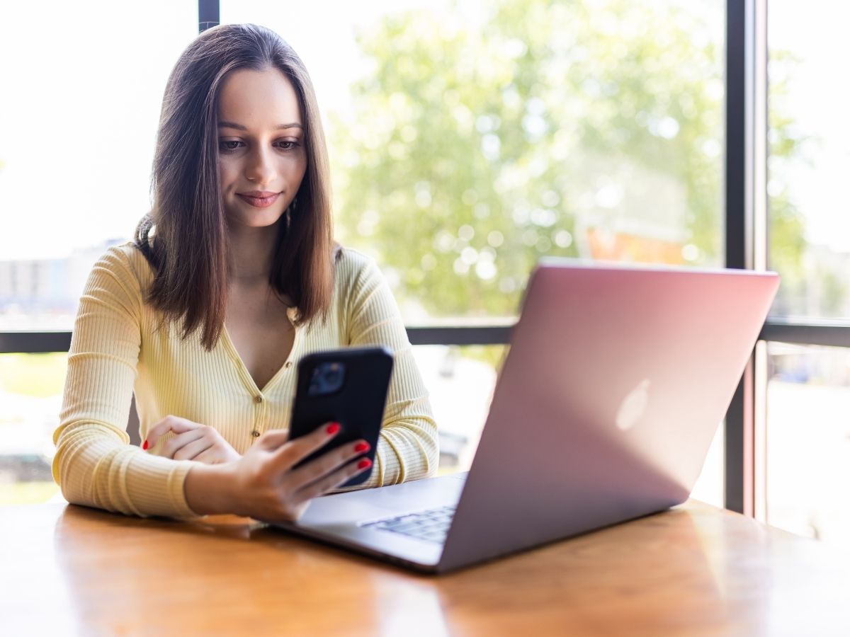 A young woman at a table with her laptop, on her phone about a course
